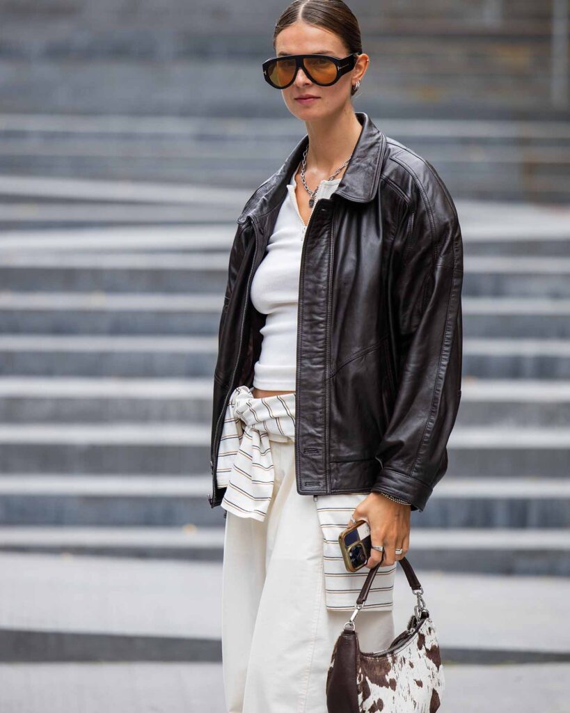 COPENHAGEN, DENMARK - AUGUST 07: Jacqueline Zelwis wears brown leather jacket, sunglasses, creme white pants, shirt, cow print bag outside Deadwood during Copenhagen Fashion Week day four on August 07, 2025 in Copenhagen, Denmark.